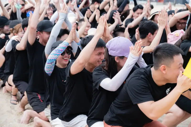 A crowd of BSS team members in black T-shirts sitting on a beach with their hands raised
