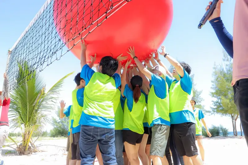 BSS team members lifting a giant red ball over a volleyball net on a sandy beach during a team game
